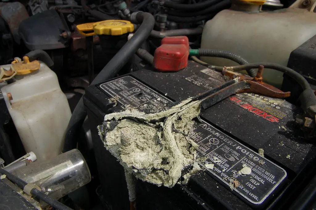 Car battery with white corrosion around the terminals in a garage, with gloves and cleanup supplies nearby