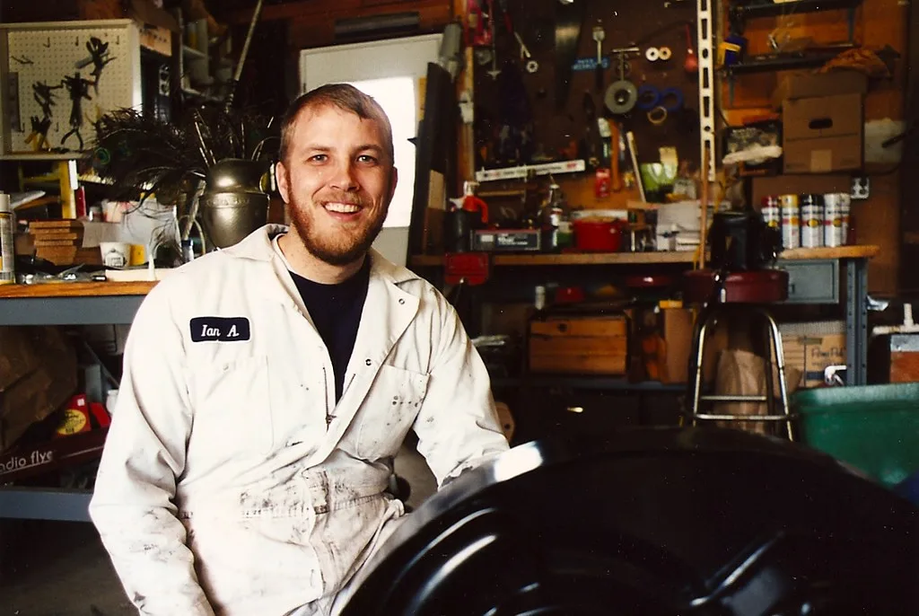 Gloved hands test a car battery terminal with a multimeter and corrosion nearby