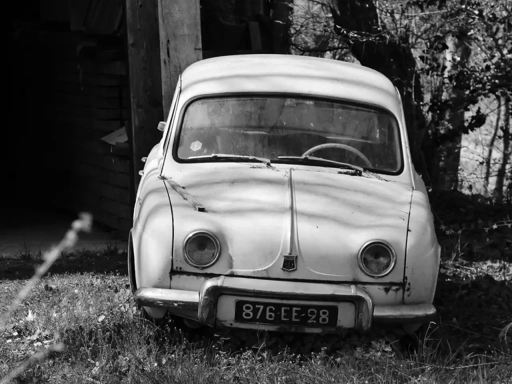 Older-car owner and mechanic inspecting an open engine bay beside a sedan in daylight