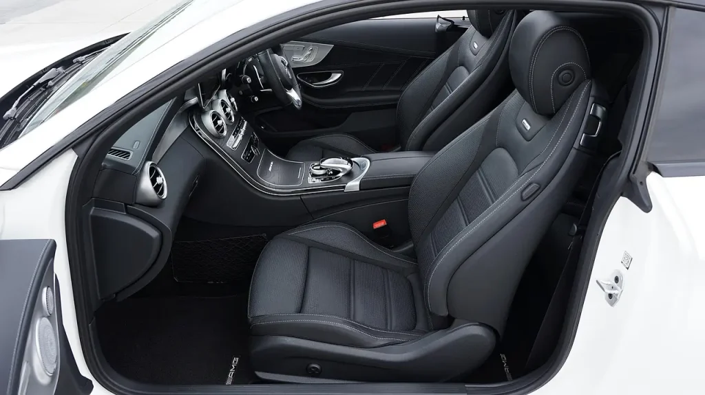 Person wiping a beige leather car seat with a microfiber cloth beside a bowl of mild soap solution