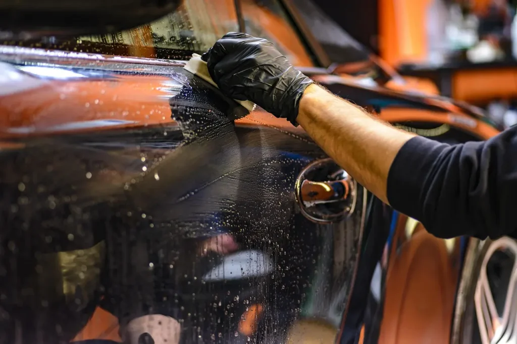 Person washing a white sedan outdoors with bucket, mitt, and hose in natural light